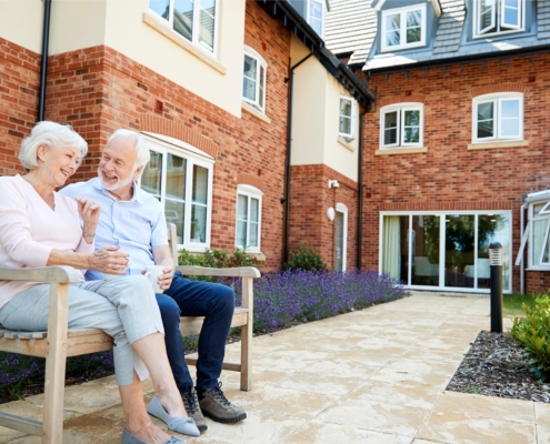 Image of two older adults sitting on a bench outside of an assisted living facility.